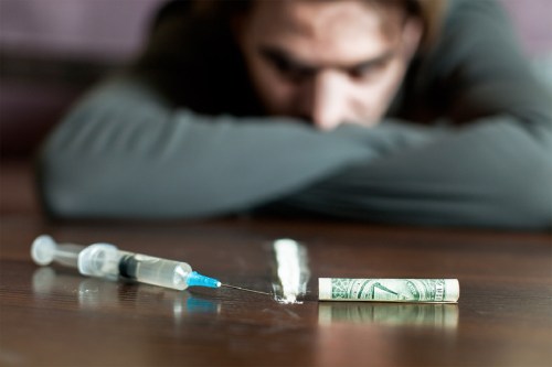 Man with arms folded on table staring at drug paraphilia including a needle and rolled up dollar bill