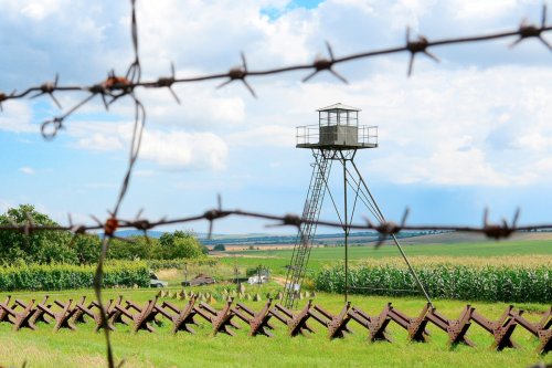 A watchtower surrounded by barbed wire with a grassy field and cornfield in the background under a cloudy sky.