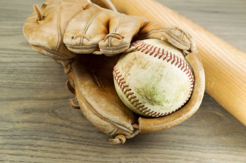Closeup horizontal photo of an old dirty baseball inside of heavily used glove and wooden bat in background on rustic wood