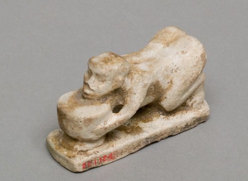 A small ancient sculpture depicting a figure leaning over a circular object, resembling a bowl or vessel, in a beige stone material.